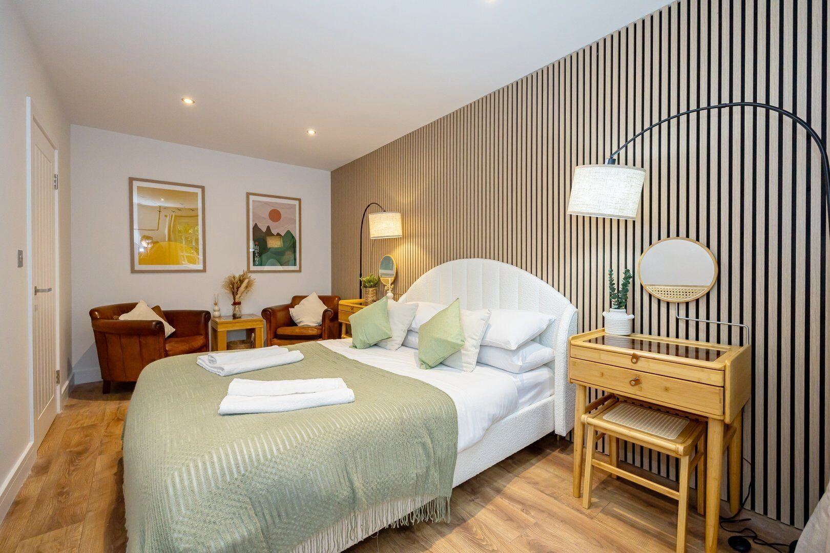 A modern hotel room with a large bed, a wooden nightstand, a desk with a chair, and a wall-mounted mirror.