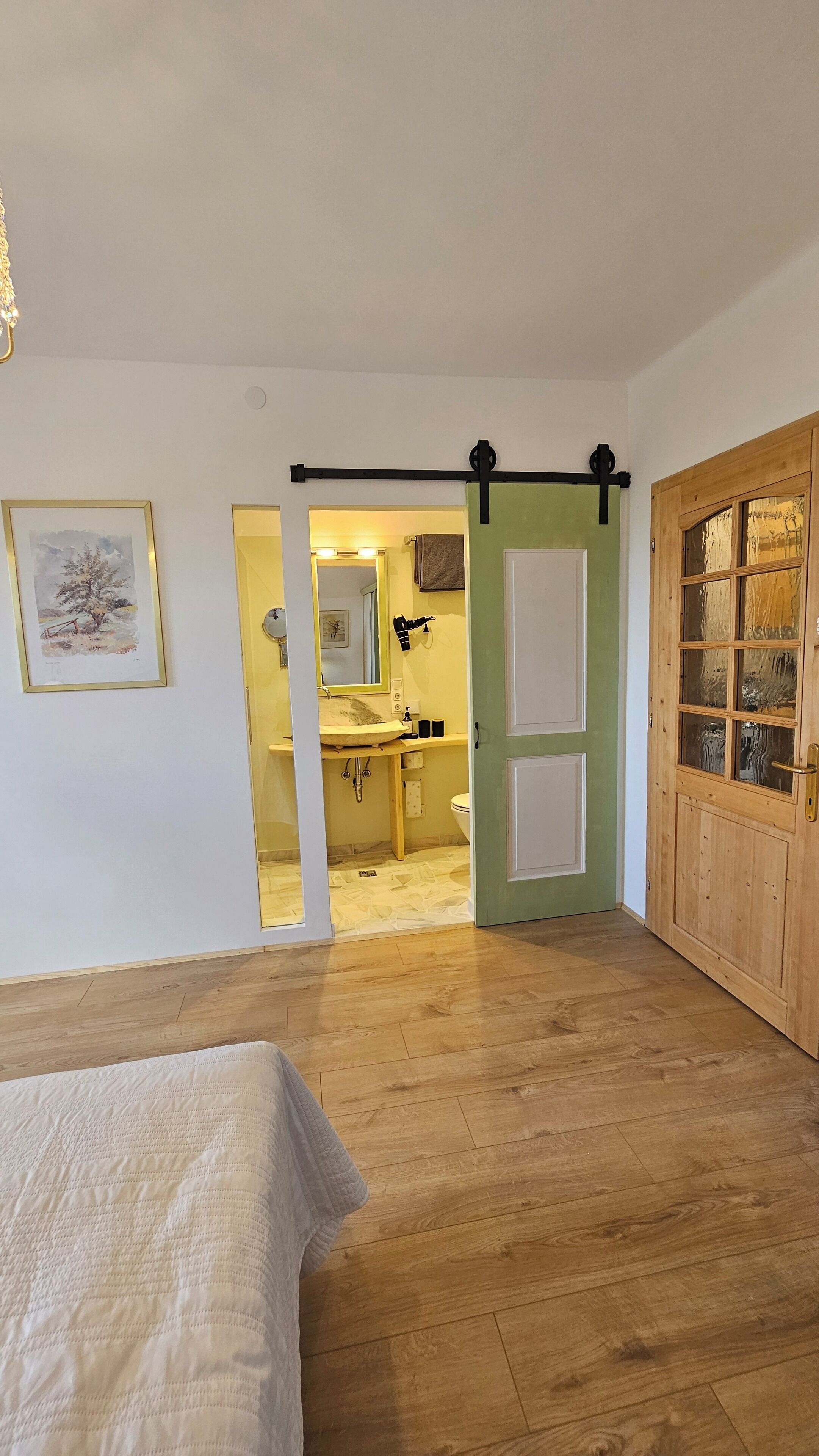 Un dormitorio con piso de madera, una cama, una puerta corredera y vistas a un baño con lavamanos y espejo.