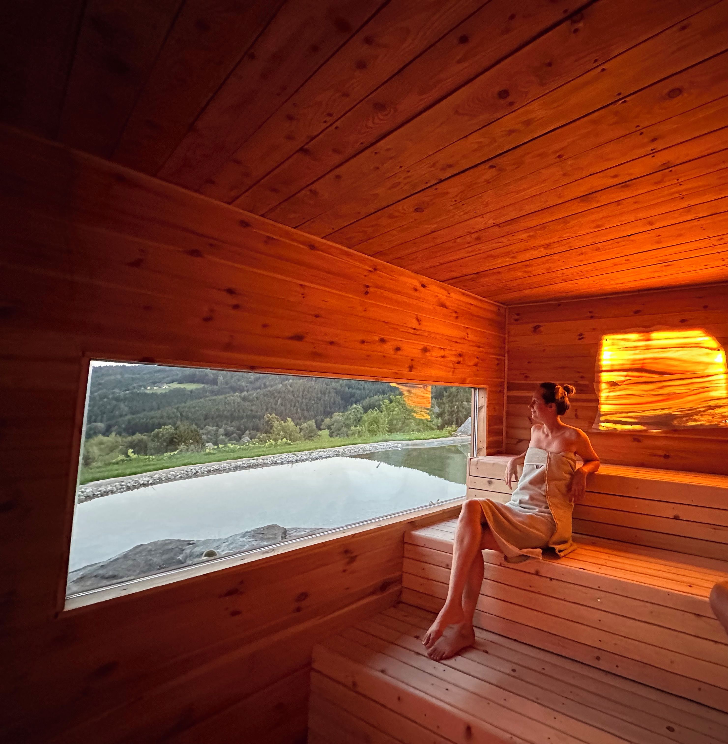 Una cabaña de madera con sauna, un ventanal grande y vistas a un lago y bosque.