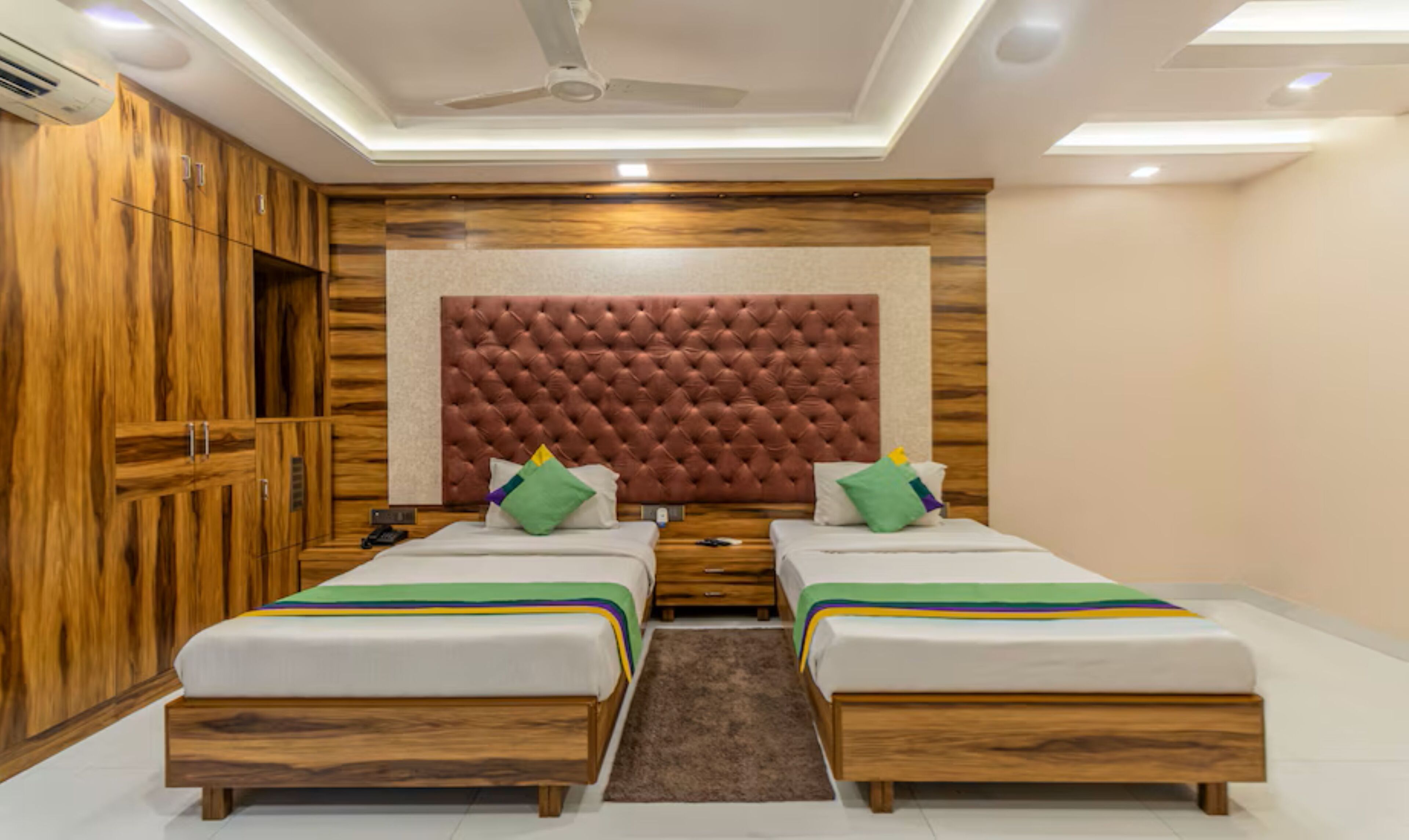 A bedroom with two single beds, a tufted headboard, wooden wardrobe, and a ceiling fan.