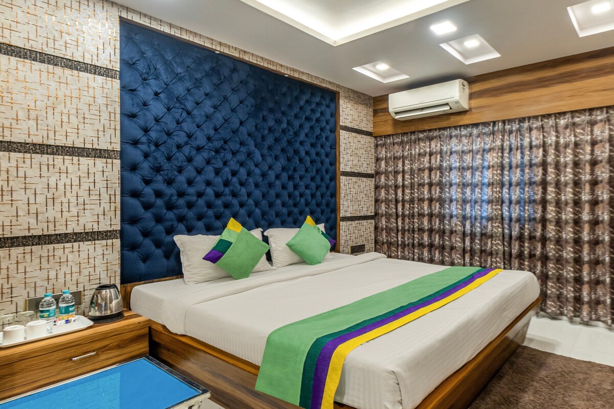 Une chambre d’hôtel moderne avec une tête de lit capitonnée, un lit recouvert d’une couvre-lit à rayures vertes et jaunes, une table de chevet en bois avec une bouilloire, et un miroir avec un cadre bleu.