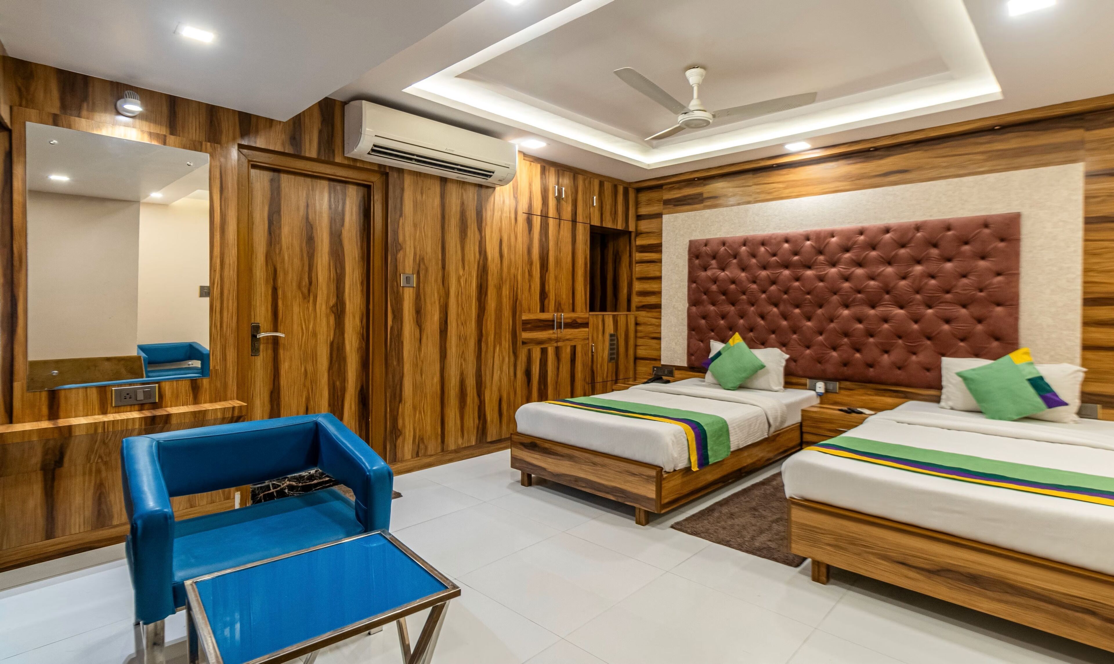A hotel room with two beds, a blue chair, a mirror, wooden wardrobe, ceiling fan, and an air conditioning unit.