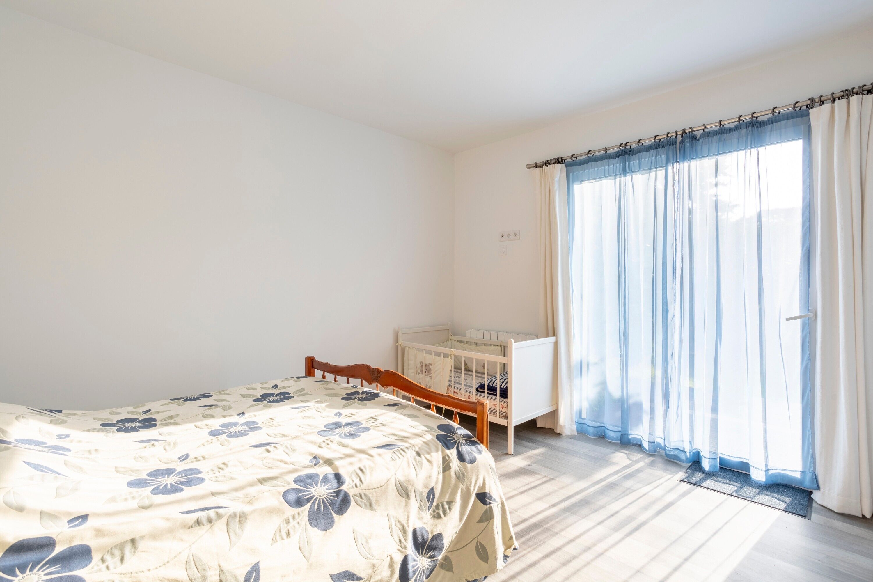 A bedroom with a bed, a crib, a window with curtains, and a door.