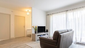 A living room with a sofa, a TV on a stand, a plant, and a window with curtains. - Holiday Home 'Maison Avec Terrasse Et Jardin' with Private Terrace, Private Garden and Wi-Fi (Saint-Yvi)