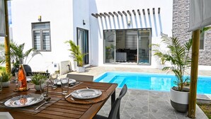 Et spiseområde ved poolen med et træbord dækket til to personer, en pool og et moderne hus i baggrunden.
