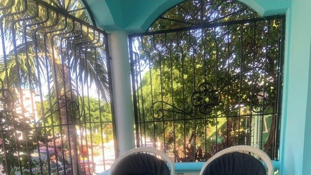 Ein überdachter Balkon mit zwei Korbstühlen und einem kleinen Tisch, umgeben von einem weißen Geländer und einem Metallrost.