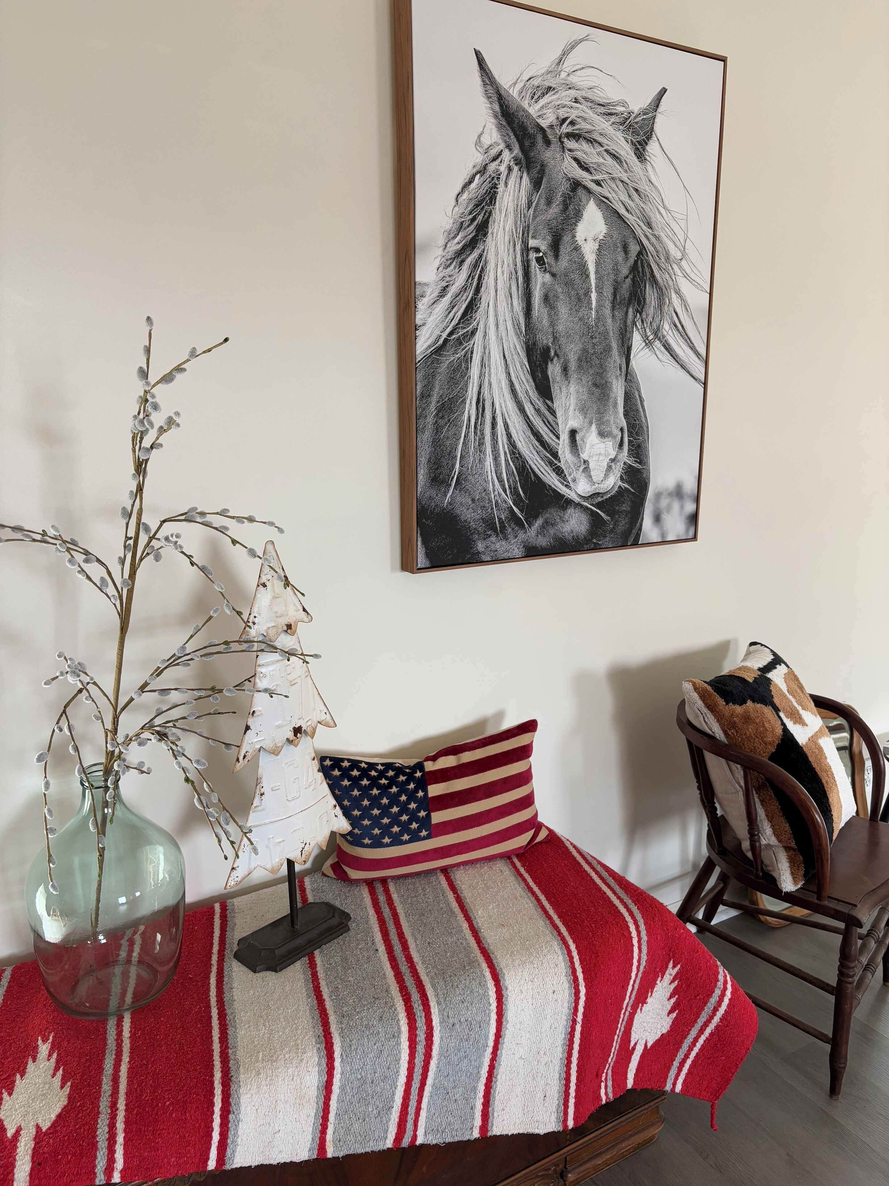 Una habitación con un retrato enmarcado de un caballo, un florero con ramas, una mesa con mantel a rayas y una silla con un cojín estampado.