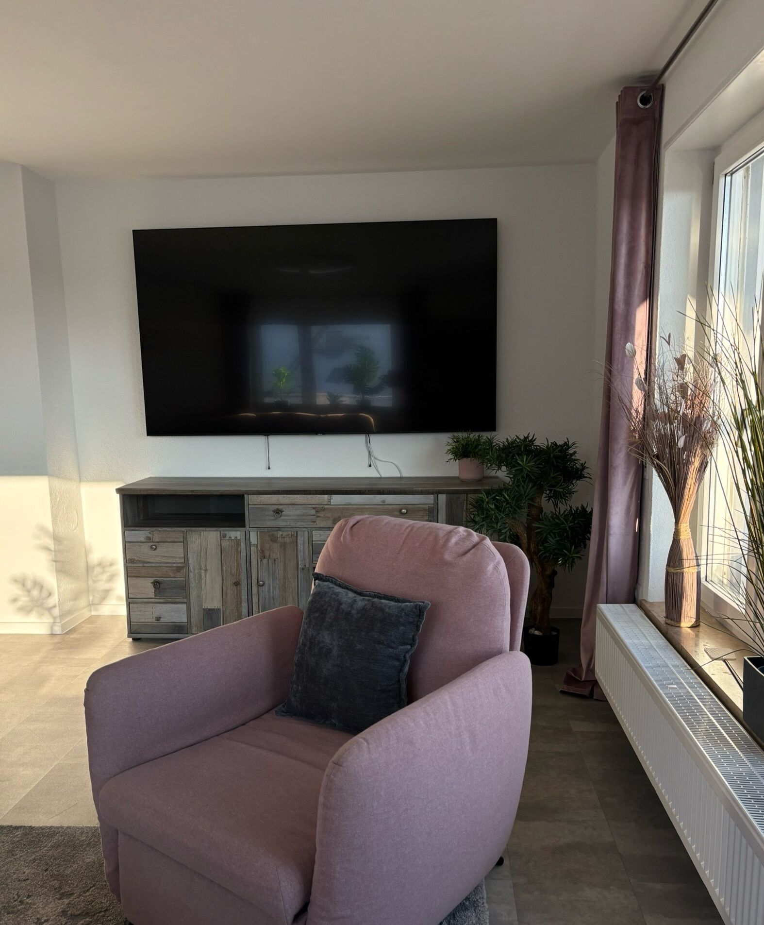 Een woonkamer met een flatscreen-tv aan de muur, een roze fauteuil met kussen, een houten kastje onder de tv en een raam met gordijnen.
