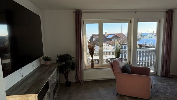 A living room with a flat-screen TV, a sofa, a potted plant, and a view of buildings through large windows. - Ferienwohnung am Weiher – Ihr perfektes Urlaubsdomizil für die ganze Familie! (Denklingen)
