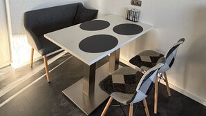 A modern dining area with a white table, black circular placemats, a grey sofa, and a patterned chair. - Ferienwohnung am Weiher – Ihr perfektes Urlaubsdomizil für die ganze Familie! (Denklingen)