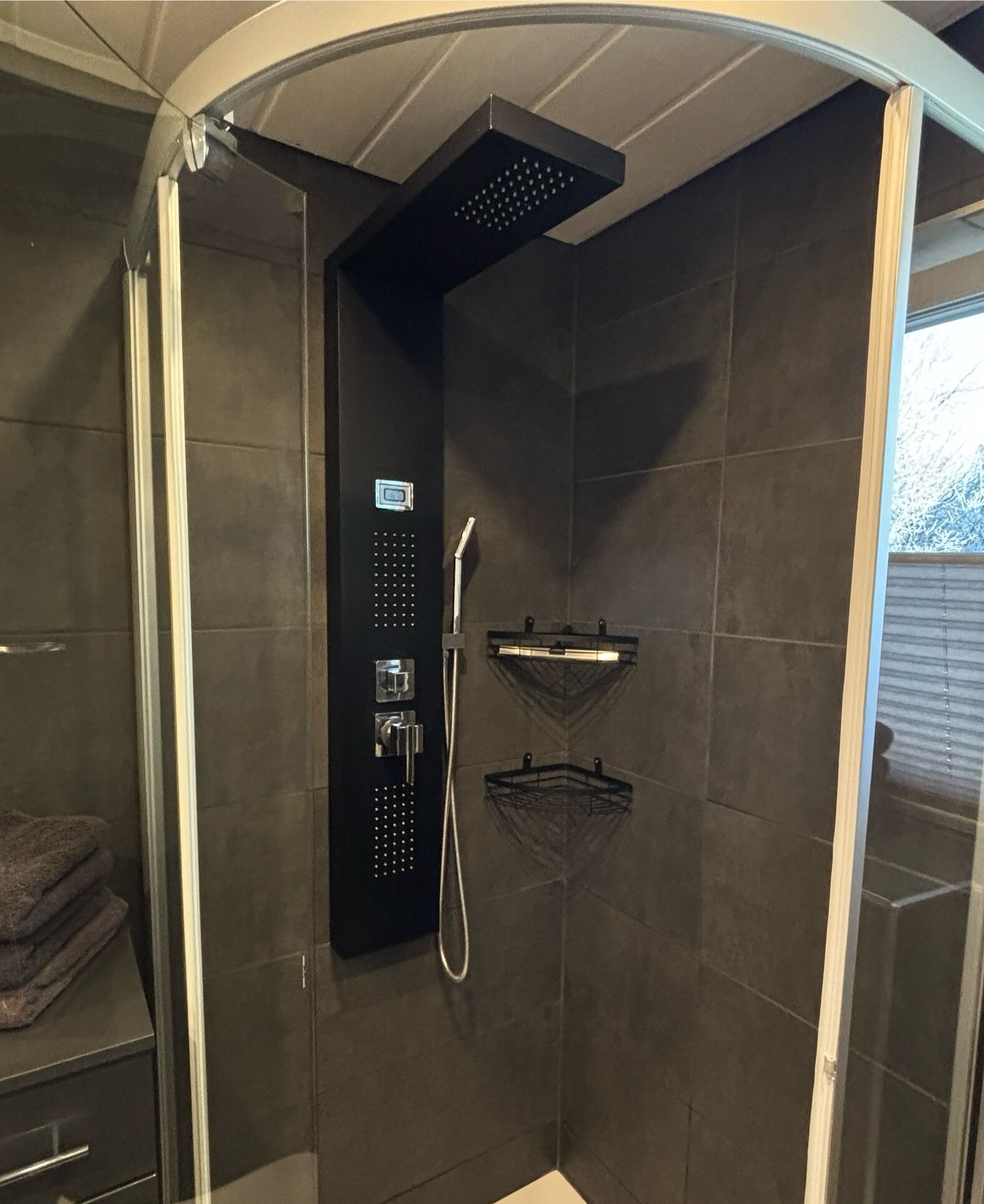 A walk-in shower with a control panel, a handheld showerhead, and a shelf.