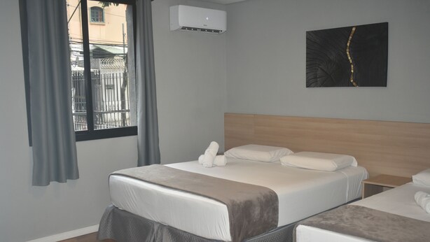 A hotel room with two beds, a wooden headboard, a gray wall, an air conditioning unit, and a window with curtains. - Unity Hotel Paulista (São Paulo)