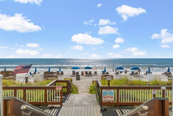 A beachside walkway leading to beach chairs and umbrellas, with an American flag and caution signs.