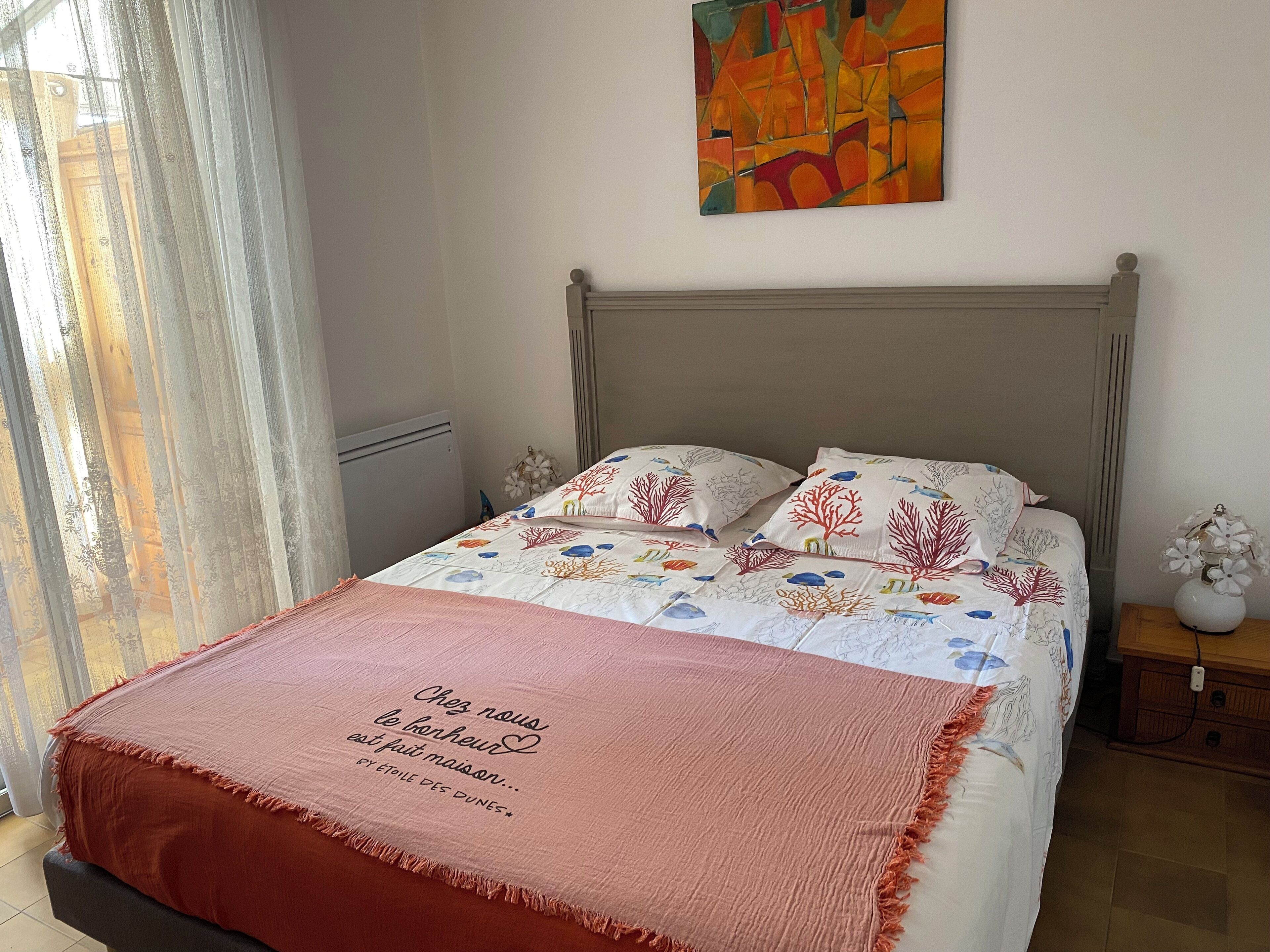 Una cama bien hecha con un cojín decorativo y una cita, una mesita de noche con un florero, y un cuadro abstracto y colorido en la pared.