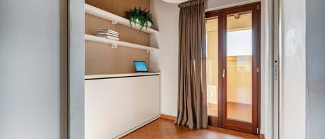 A room with a white desk, shelves with books and a plant, a laptop, a large window with curtains, and a wooden ceiling.