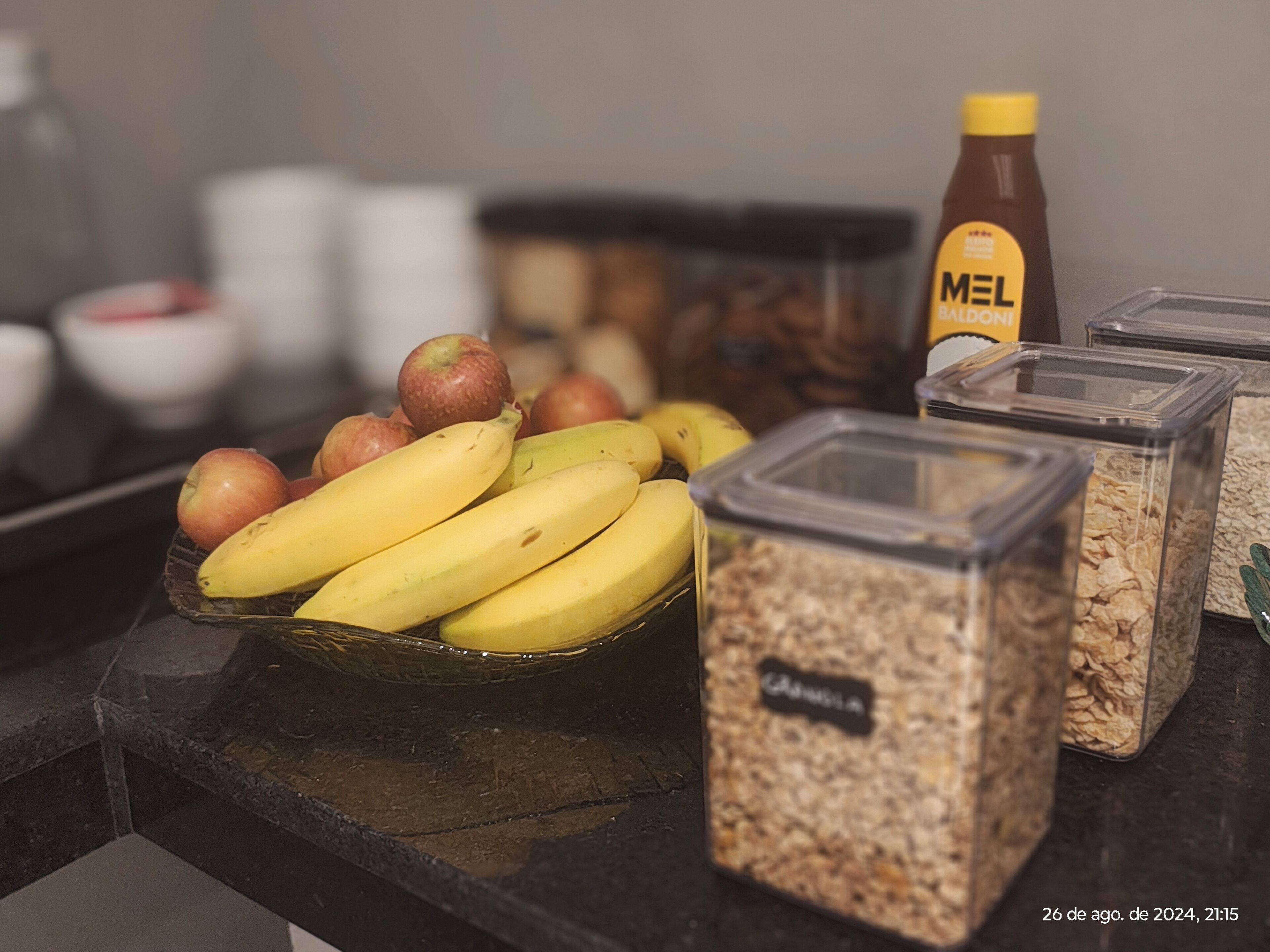 A breakfast bar with bananas, apples, cereal, and a bottle of honey.