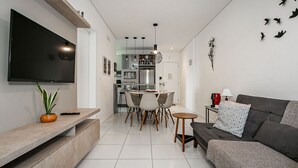 A modern living room with a flat-screen TV, a sofa, a dining area with chairs, and a kitchen in the background.