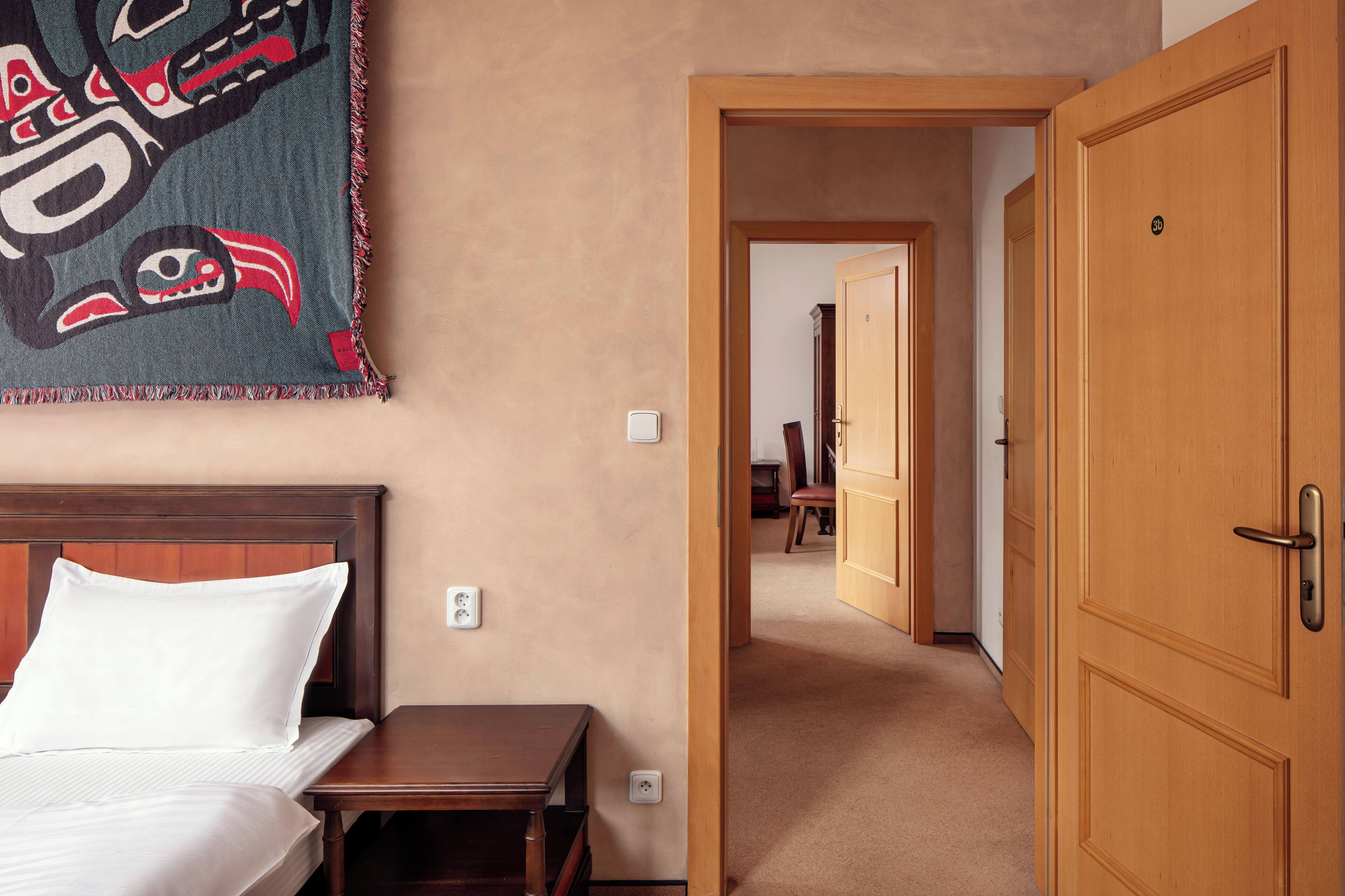 Una camera d'albergo con un letto, un tavolino, un arazzo a parete e una porta in legno.