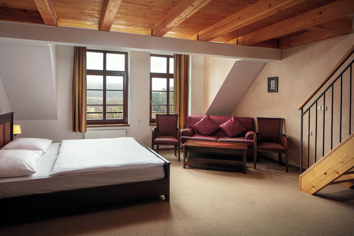 A bedroom with a bed, a sofa, a chair, a wooden staircase, and a window with curtains.