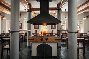 Un restaurante con un horno de leña central, suelo de piedra y vigas de madera. Mesas y sillas están dispuestas alrededor del horno.