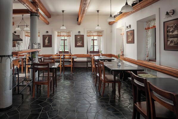 A restaurant with wooden tables and chairs, stone flooring, and hanging pendant lights. - Hotel Javor (Adrspach)