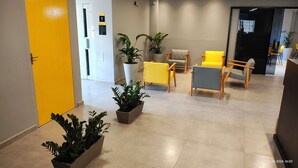 Un hall d’entrée moderne avec une porte jaune, plusieurs coins salon avec des fauteuils jaunes et gris, et des plantes en pot.