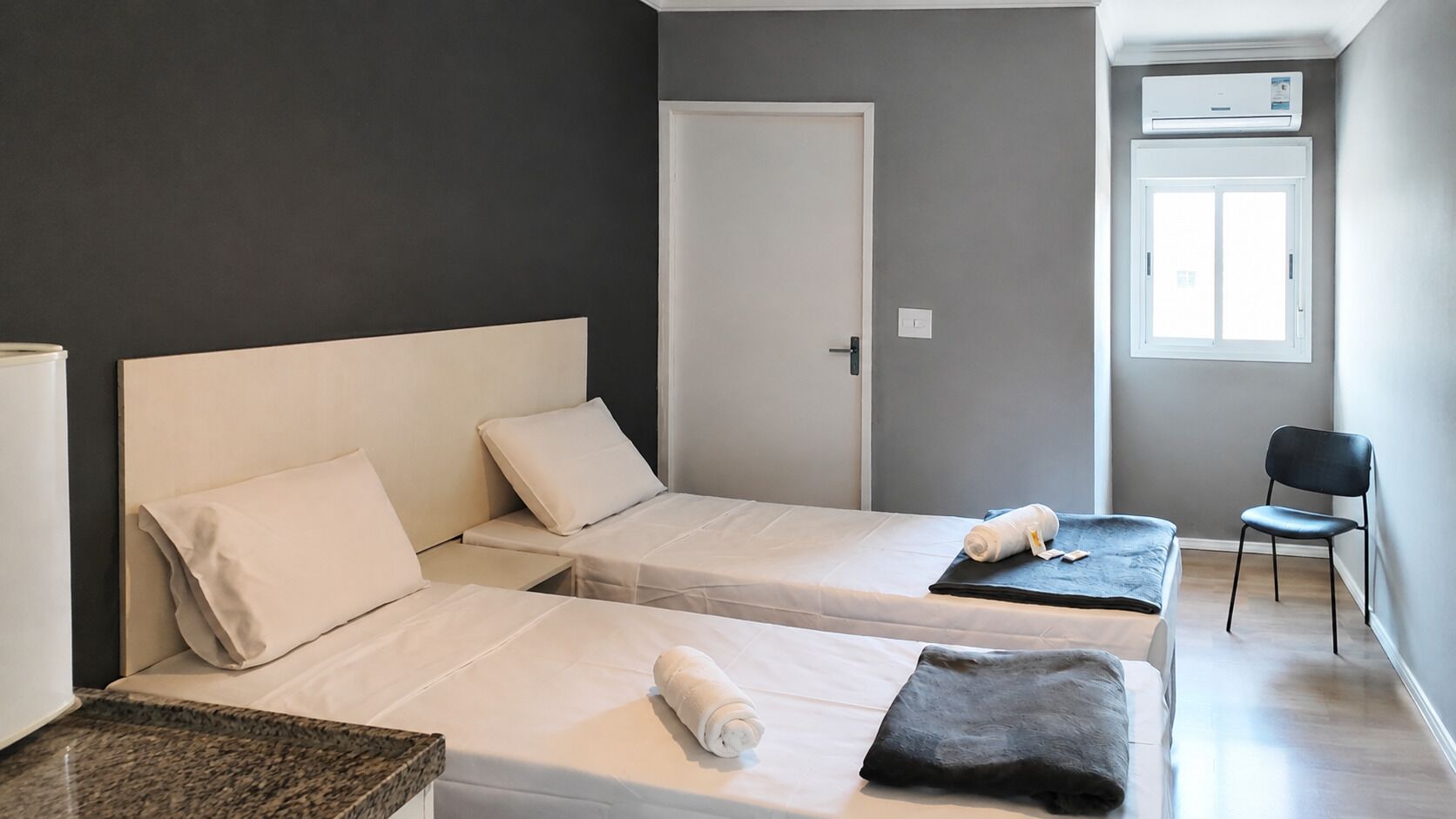 A hotel room with two single beds, a small table, a chair, a window, and an air conditioning unit.