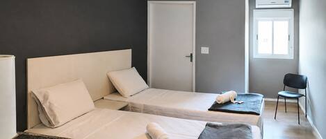A hotel room with two single beds, a small table, a chair, a window, and an air conditioning unit.