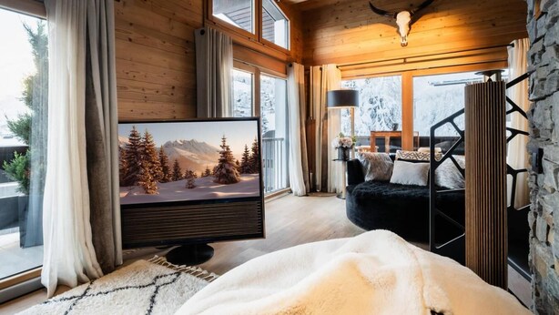 A cozy room with a large flat-screen TV, wooden walls, a stone accent wall, and a view of snow-covered mountains.