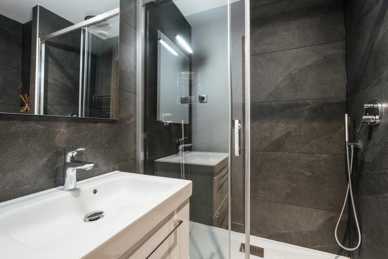 Un baño moderno con cabina de ducha acristalada, un lavabo blanco y paredes de baldosas oscuras.