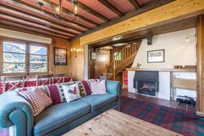 A cozy living room with a sofa, a fireplace, a wooden staircase, and a view of the mountains through large windows.