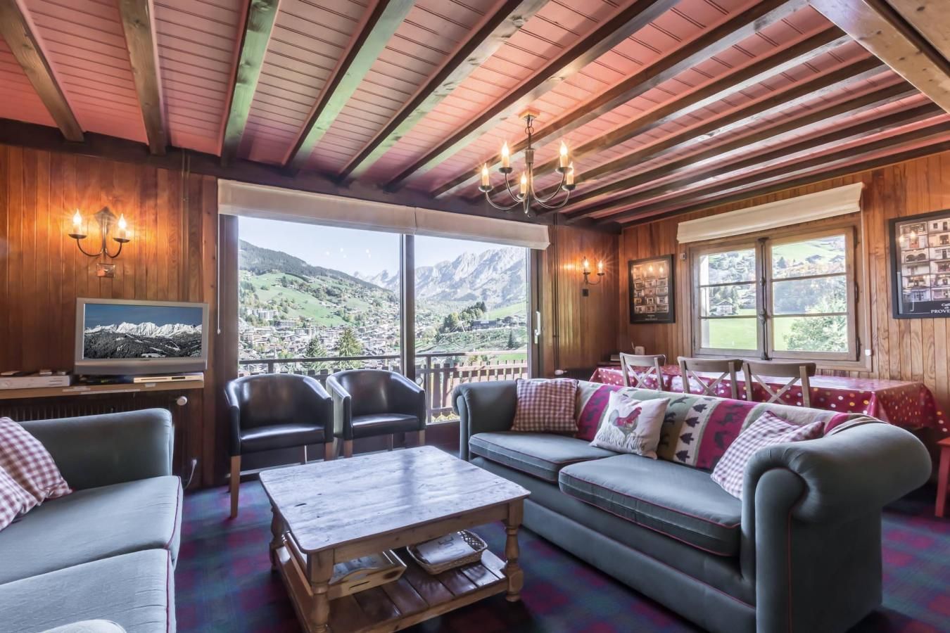 A cozy room with a wooden ceiling, a large window offering a mountain view, a television, and a dining area with a table and chairs.