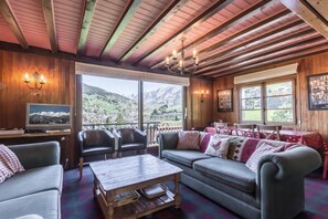 A cozy room with a wooden ceiling, a large window offering a mountain view, a television, and a dining area with a table and chairs.