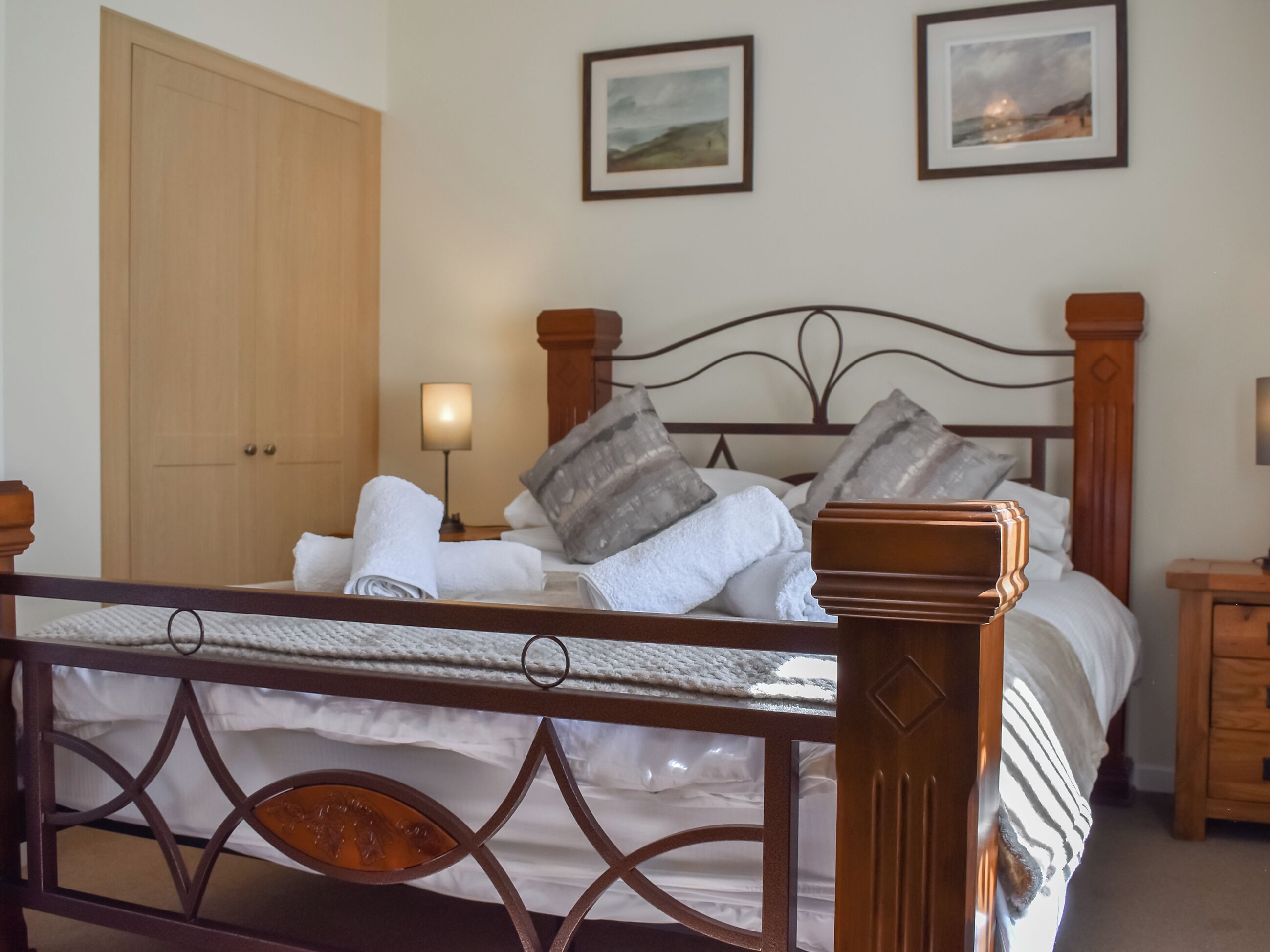 A neatly made bed with white linens and rolled towels, a wooden headboard, and two framed pictures on the wall.