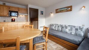 A compact kitchen with wooden cabinets, a microwave, and a coffee maker. There is a round wooden table with chairs and a grey patterned sofa.