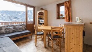 A cozy room with a wooden dining table, chairs, a sofa with patterned cushions, a wooden cabinet, and a window with a view of snow-covered mountains.