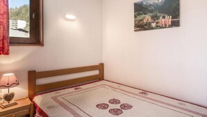 A bedroom with a wooden bed, a bedside table with a lamp, a window with a view of buildings, and a wall-mounted picture of a landscape.
