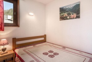 A bedroom with a wooden bed, a bedside table with a lamp, a window with a view of buildings, and a wall-mounted picture of a landscape.