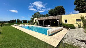 A pool area with lounge chairs, a covered seating area, and a building with a tiled roof. - Spacious cottage with air conditioning, wood fire & panoramic views - pool shared (Paulhiac)
