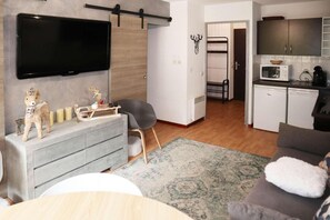 A modern living room with a grey sofa, a flat-screen TV mounted on the wall, a small dining table, and a kitchen area with a microwave and cabinets.
