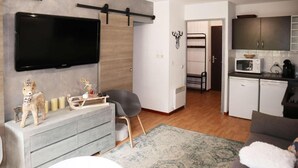 Een moderne woonkamer met een grijze bank, een flatscreen televisie aan de wand, een eettafeltje en een keuken met een magnetron en kastjes.