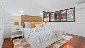 Quarto com cama grande, paredes brancas, soalho de madeira e janela com vista para árvores.