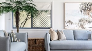 Uma sala de estar com um sofá azul-claro, uma cesta de vime, uma janela grande com vista para palmeiras e uma pintura de uma cena de praia na parede.