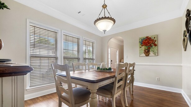 Une salle à manger avec une table en bois, des chaises blanches et un lustre. De grandes fenêtres sont équipées de stores, un tableau orne le mur et une porte donne accès à une autre pièce.