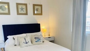 Una cama cuidadosamente tendida con un banco tapizado en azul, ropa de cama blanca y almohadones con motivos náuticos.