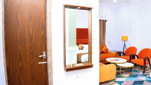 Une chambre d’hôtel avec une porte en bois, un miroir, un lit avec une literie rouge, des chaises orange, une petite table ronde et un tapis à motifs.