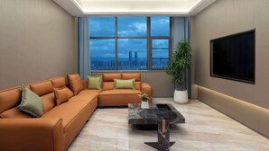A modern living room with a sectional sofa, a coffee table, a large window with a city view, and a flat-screen TV mounted on the wall. - Days Inn By Wyndham Kunming Wujiaba (Kunming)