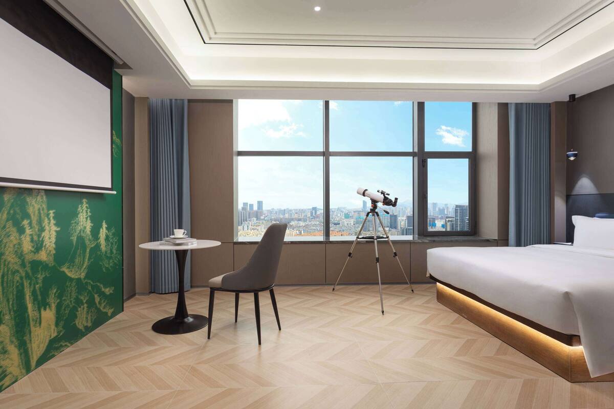 A hotel room with a large bed, a desk with a chair, a telescope, and a city view through large windows.