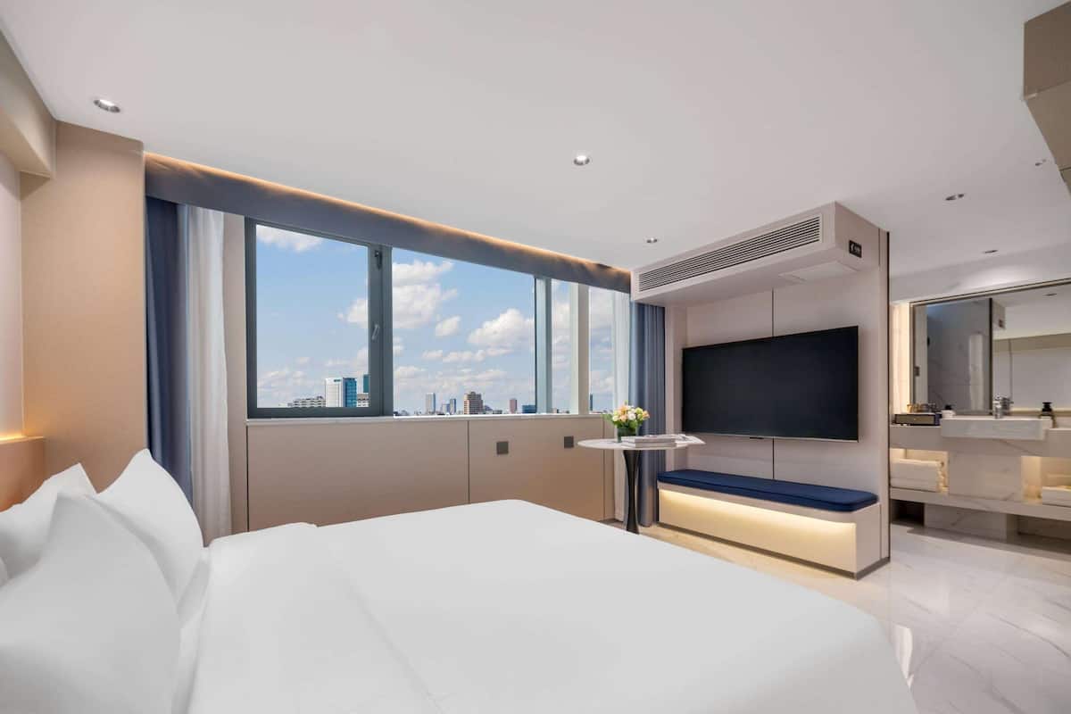 A modern hotel room with a large bed, a TV, a small table with a bench, and a city view through large windows.