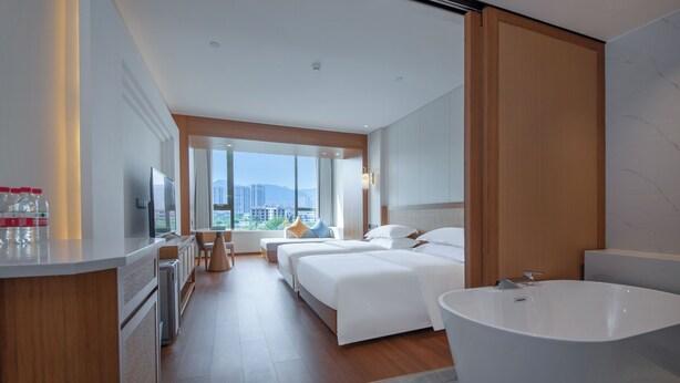 Una moderna camera d'albergo con un letto grande, un bagno separato con vasca freestanding, una scrivania con una sedia e un'ampia finestra con vista sulla città.